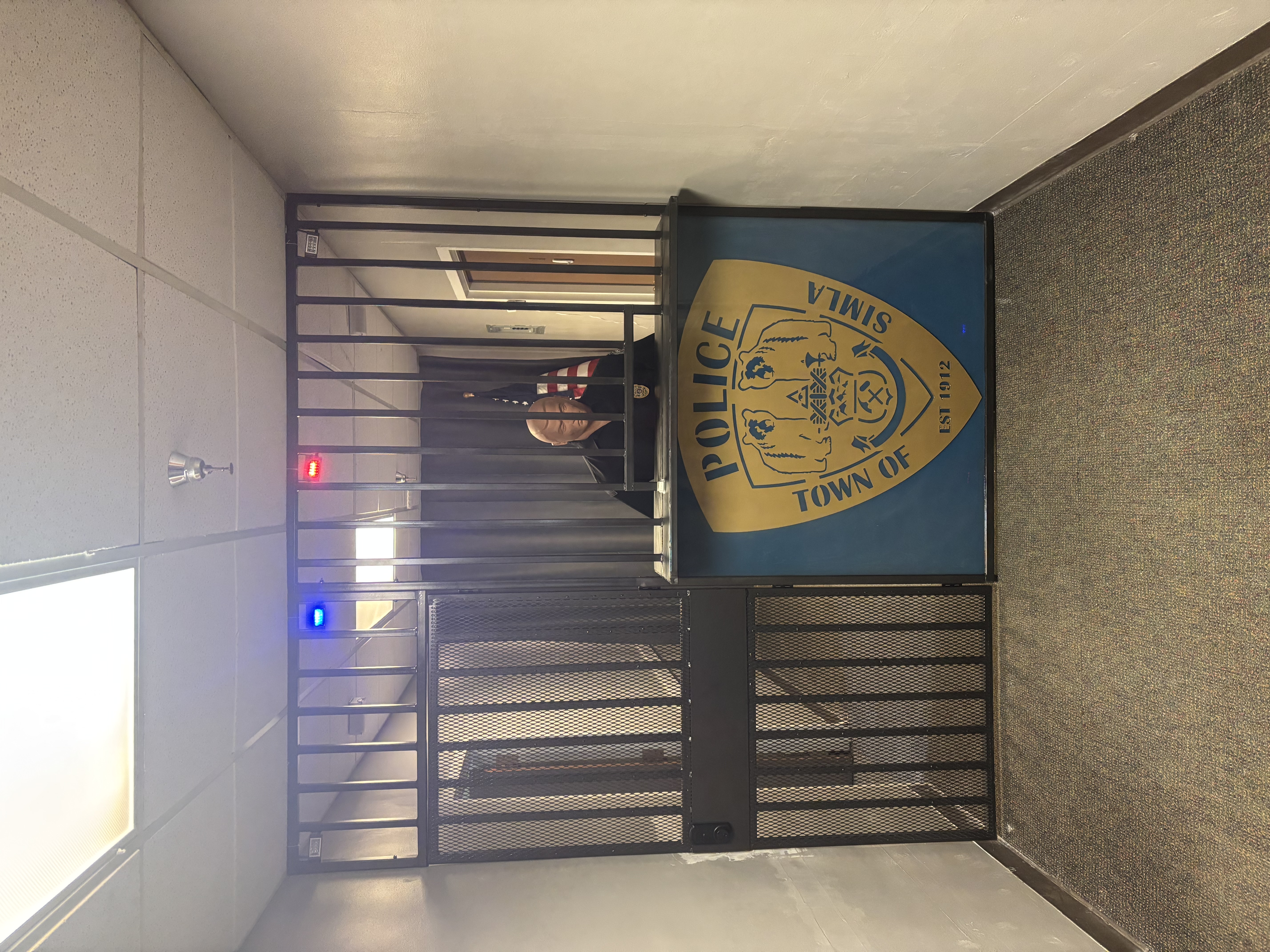 Picture of the Front entrance to the Police Department from the Hope Community Center's Grand Opening on April 5th, 2025. Black bars with a black security door. There are flashing red and blue lights as well as a male mannequin. 